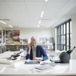 Smiling female architect talking on cell phone and using digital tablet at table in office